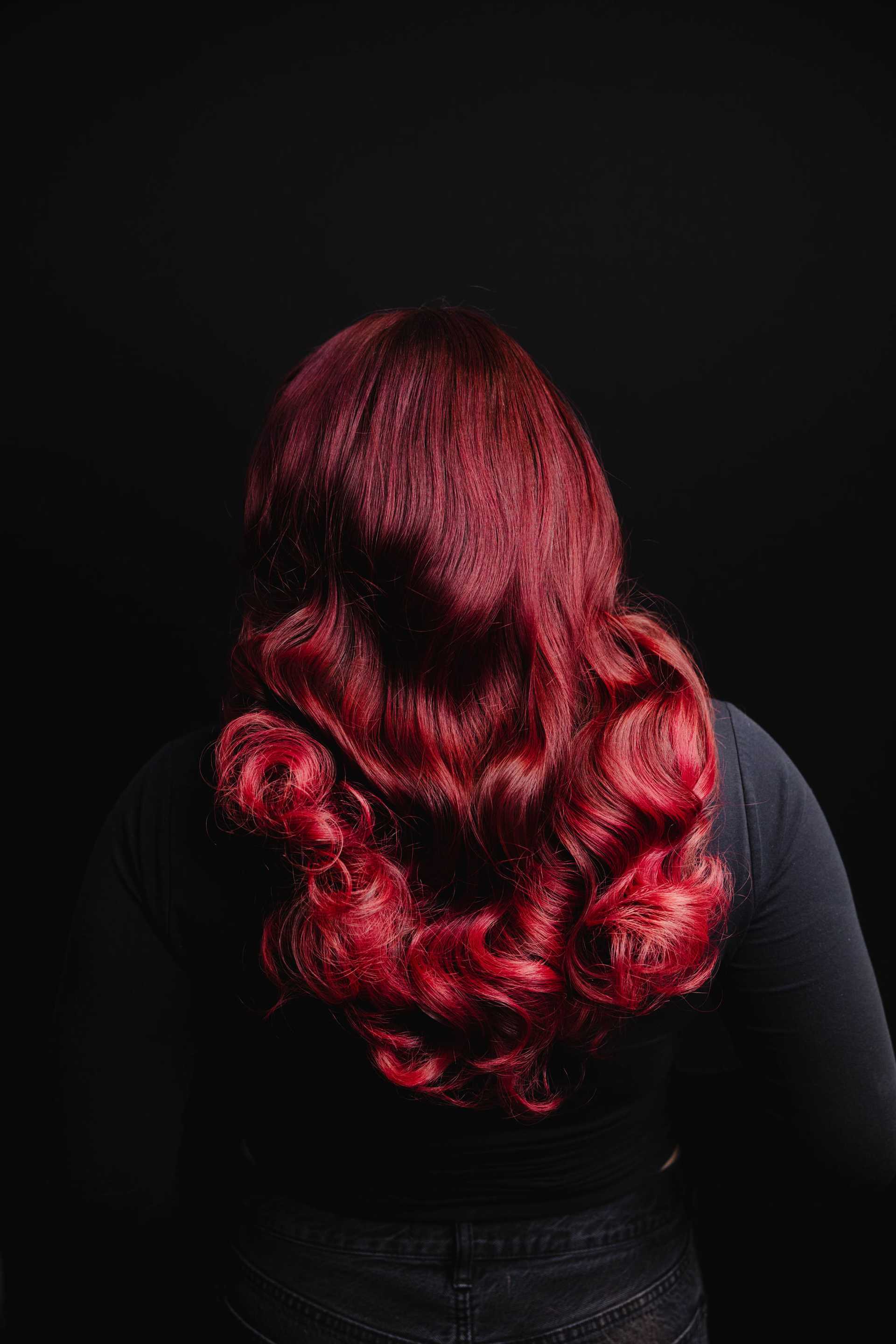 Red-haired person with long, curly hair against a dark background.