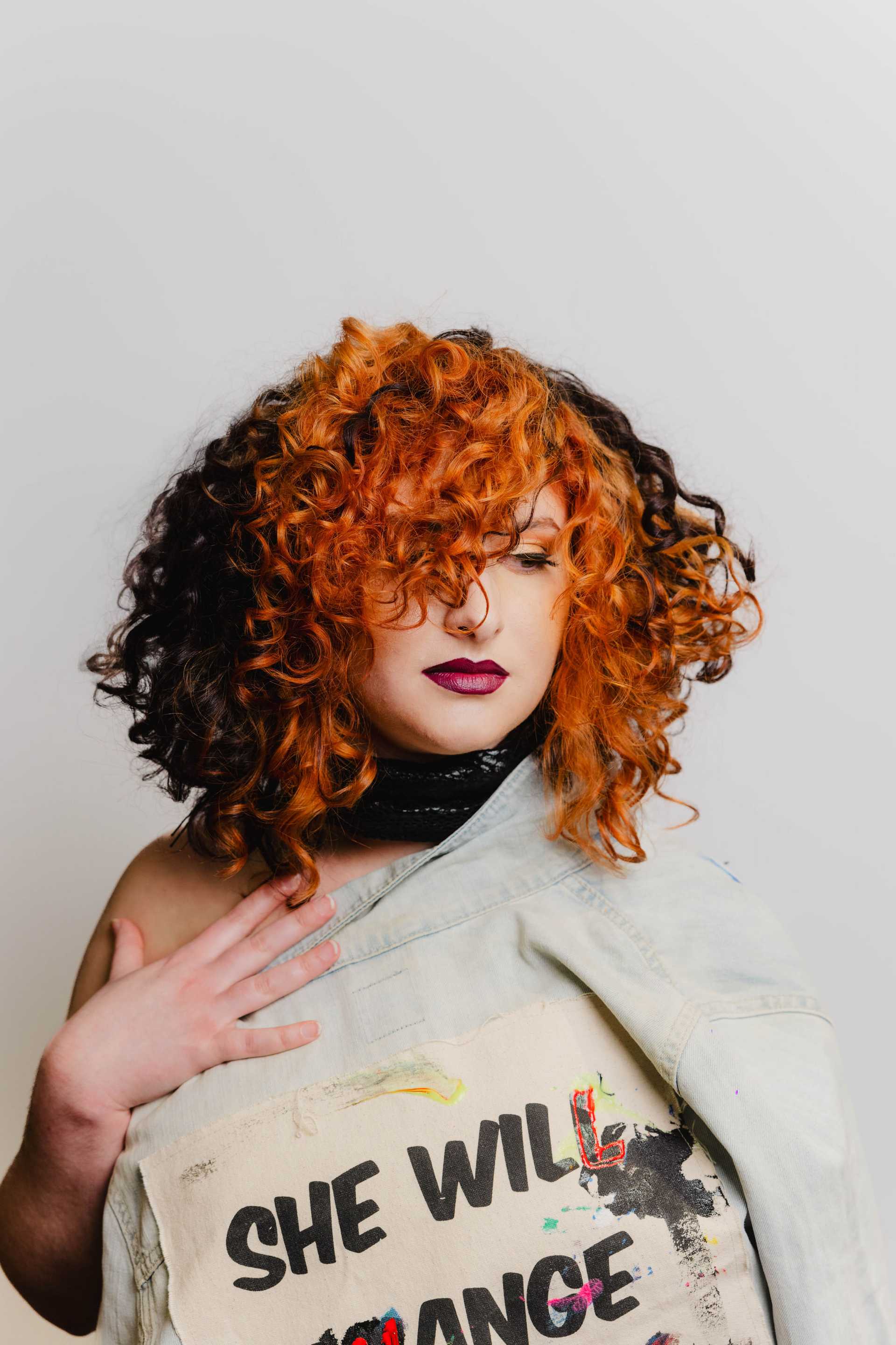 Person with curly orange hair holding jacket and wearing makeup.