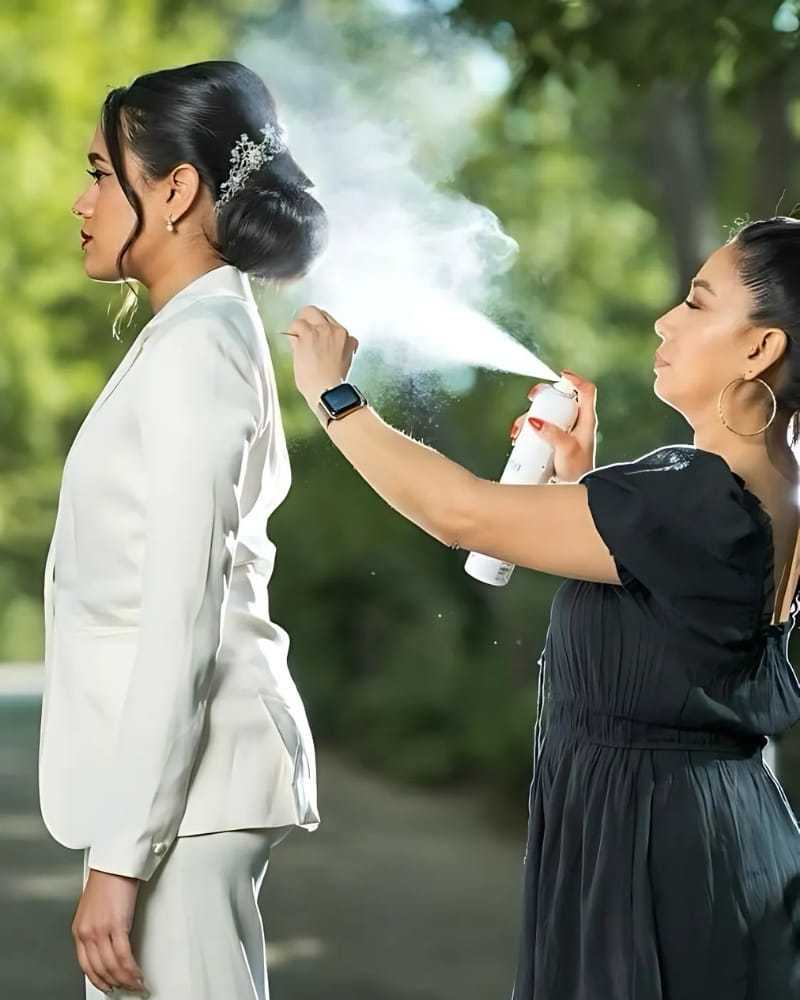 Woman in suit gets hair styled outdoors with hairspray by another person.