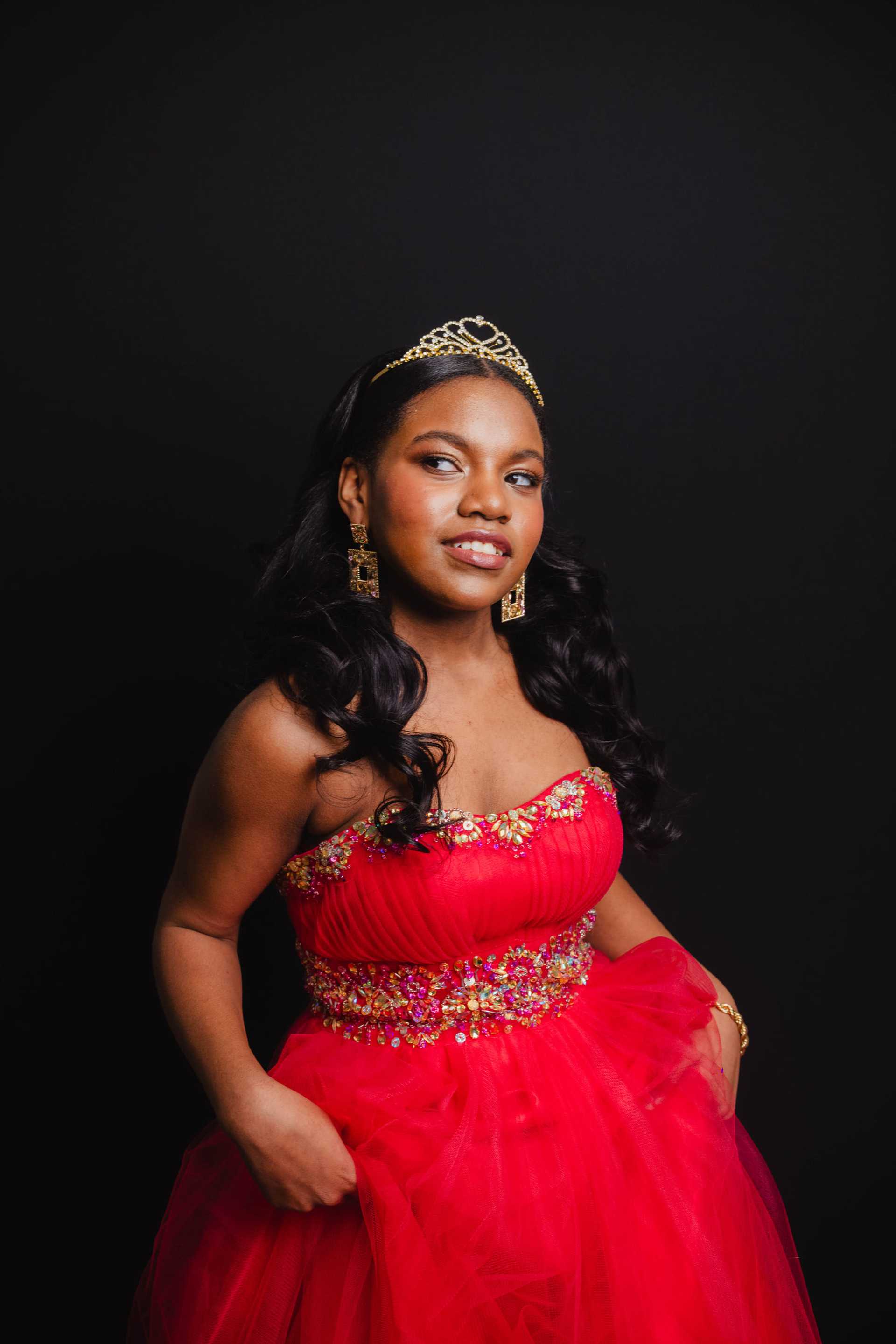 Young woman in a red gown and tiara, posing confidently against a dark background.