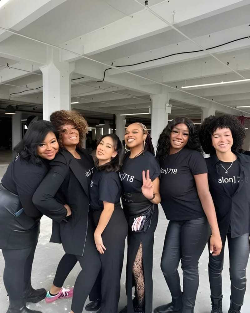 Group of six smiling people in matching black Salon 718 shirts in an industrial space.