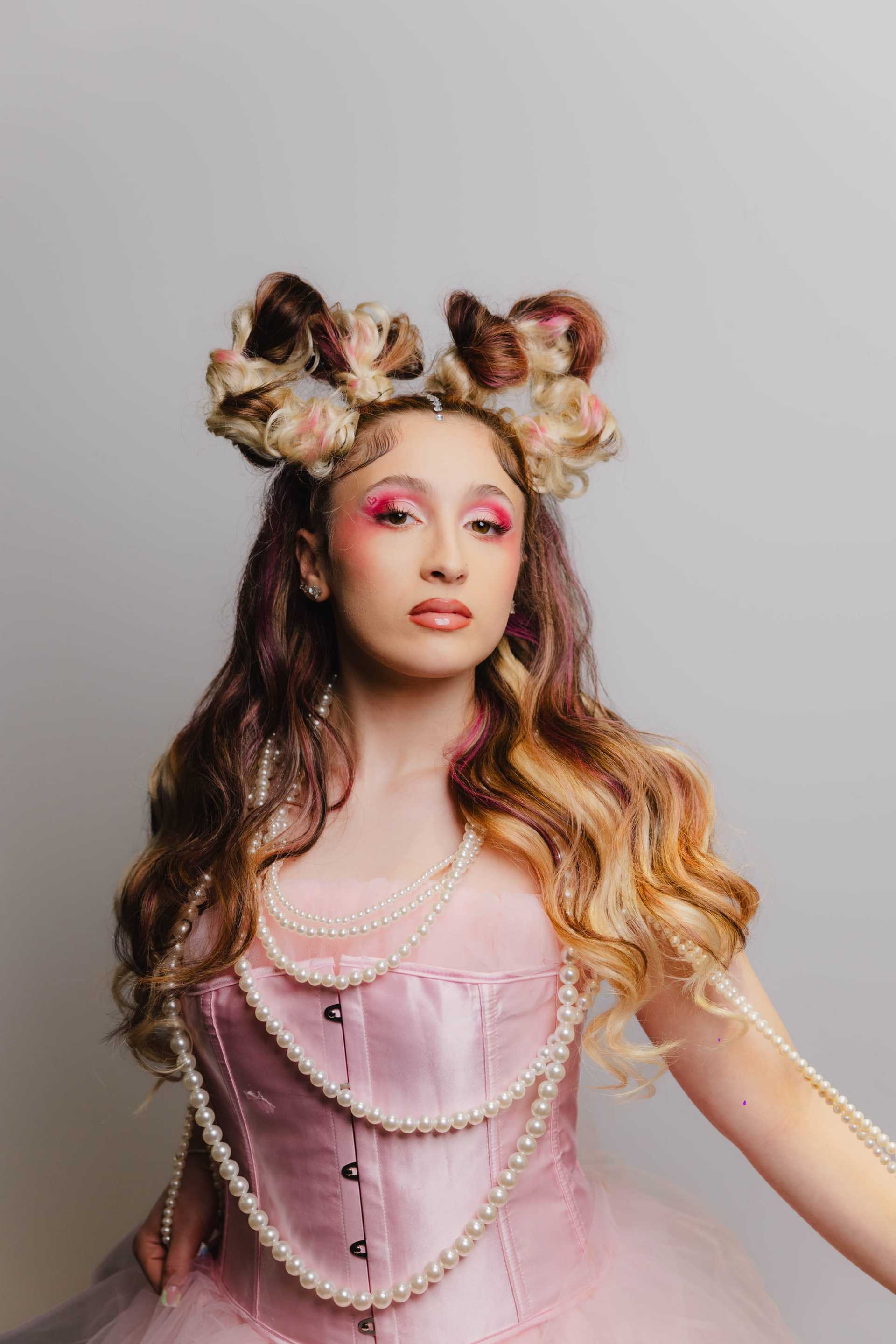 Young woman in elaborate hairstyle and pink dress with pearls, against a gray background.