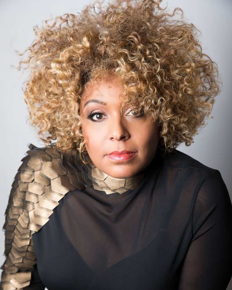 Woman with curly hair in black outfit, gold accents.